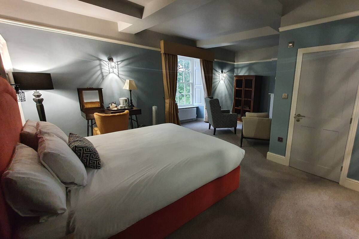A hotel room with a large bed, a desk with a lamp, a chair, and a wardrobe.