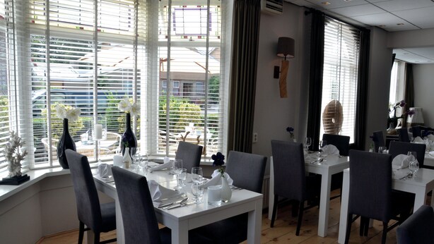 A dining area with white tables and grey chairs, large windows with blinds, and a view of the outside. - Hotel Koogerend (Den Burg)