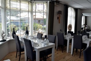 Un comedor con mesas blancas y sillas grises, ventanas amplias con persianas y vistas al exterior.