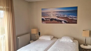 A hotel room with a double bed, two bedside tables with lamps, a large window with curtains, a radiator, and a painting of a beach scene on the wall. - Hotel Koogerend (Den Burg)