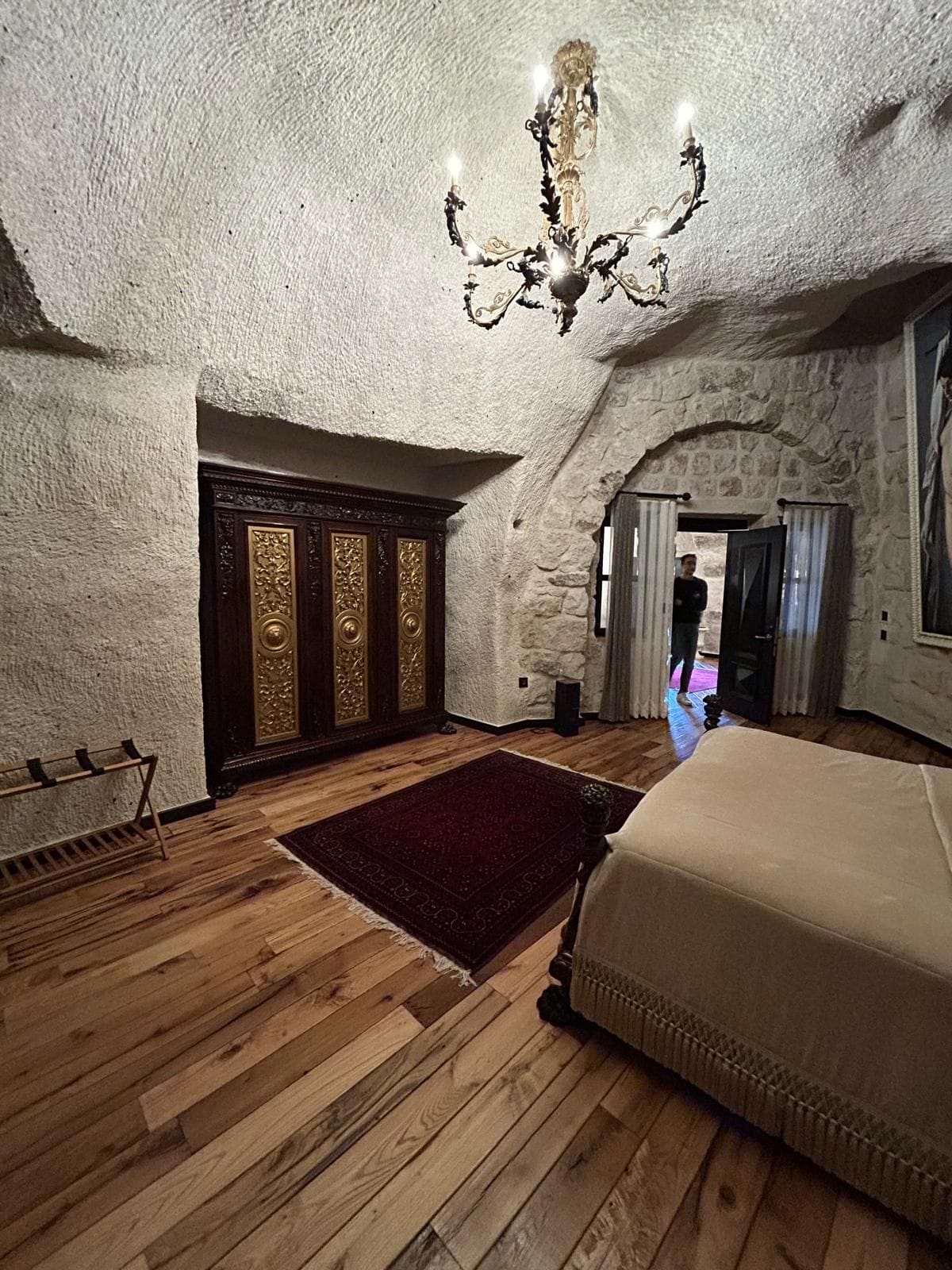A bedroom with a wooden floor, a large bed, a chandelier, and a stone wall.