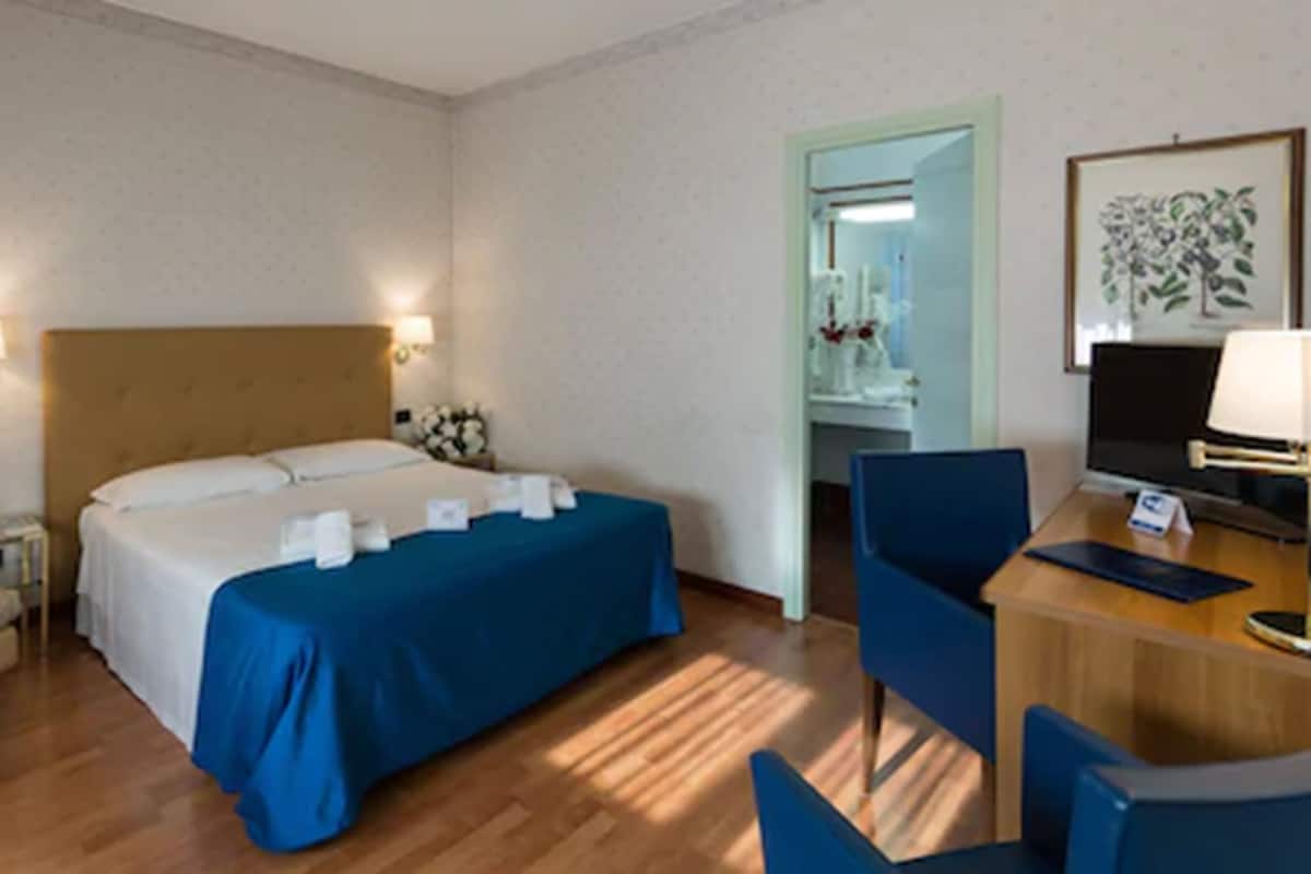 A hotel room with a bed, a desk, a chair, a sofa, and a bathroom visible through an open door.