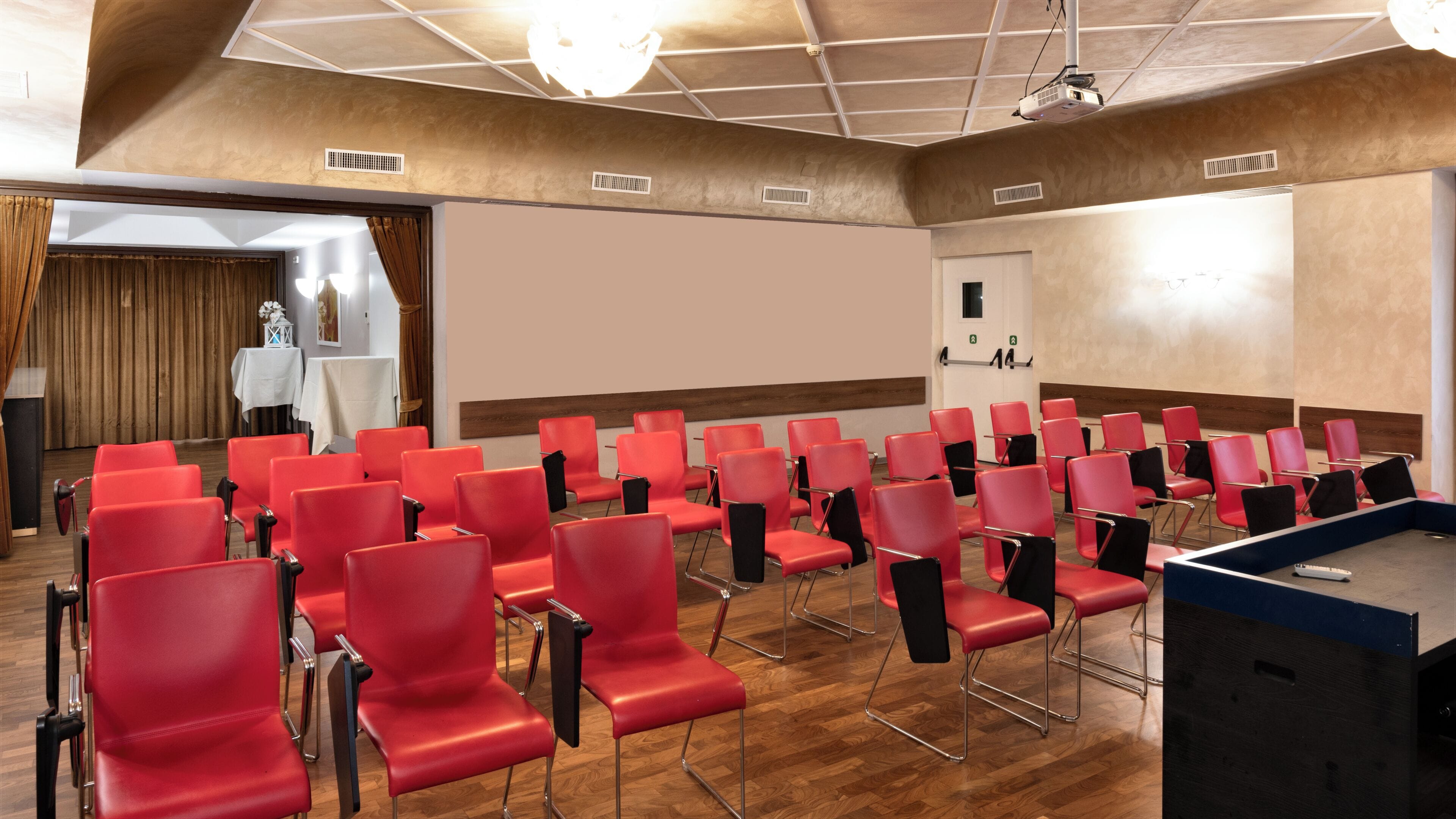 A conference room with rows of red chairs, a projector screen, and a wooden floor.