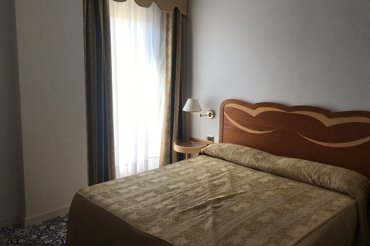 Et hotelværelse med en pænt redt seng, et sengebord med lampe, og et vindue med gardiner.