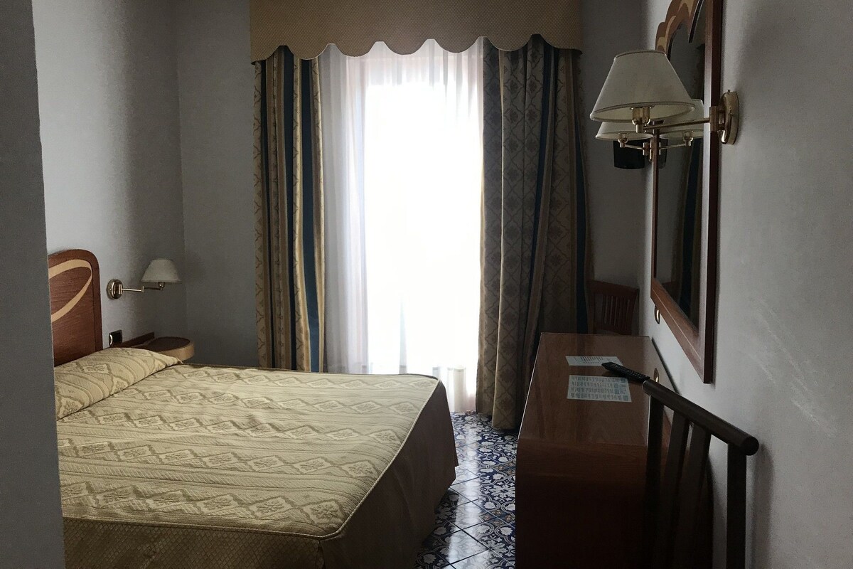 Et hotelværelse med seng, skrivebord, stol, lampe og et vindue med gardiner.