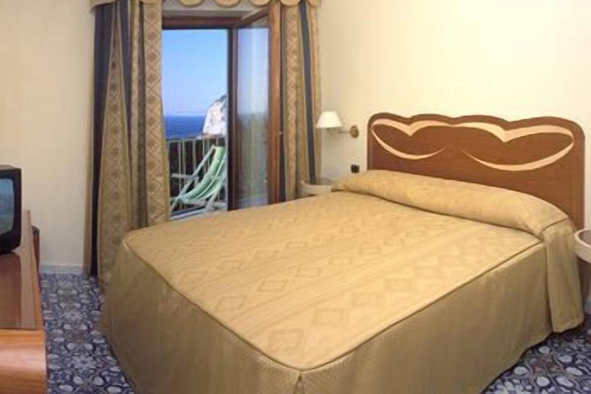 Een hotelkamer met een bed, een televisie en uitzicht op zee door een raam.