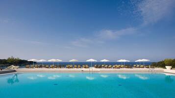 Una piscina azul cristalina con sillas de descanso y sombrillas, rodeada por un mar tranquilo y un cielo despejado.