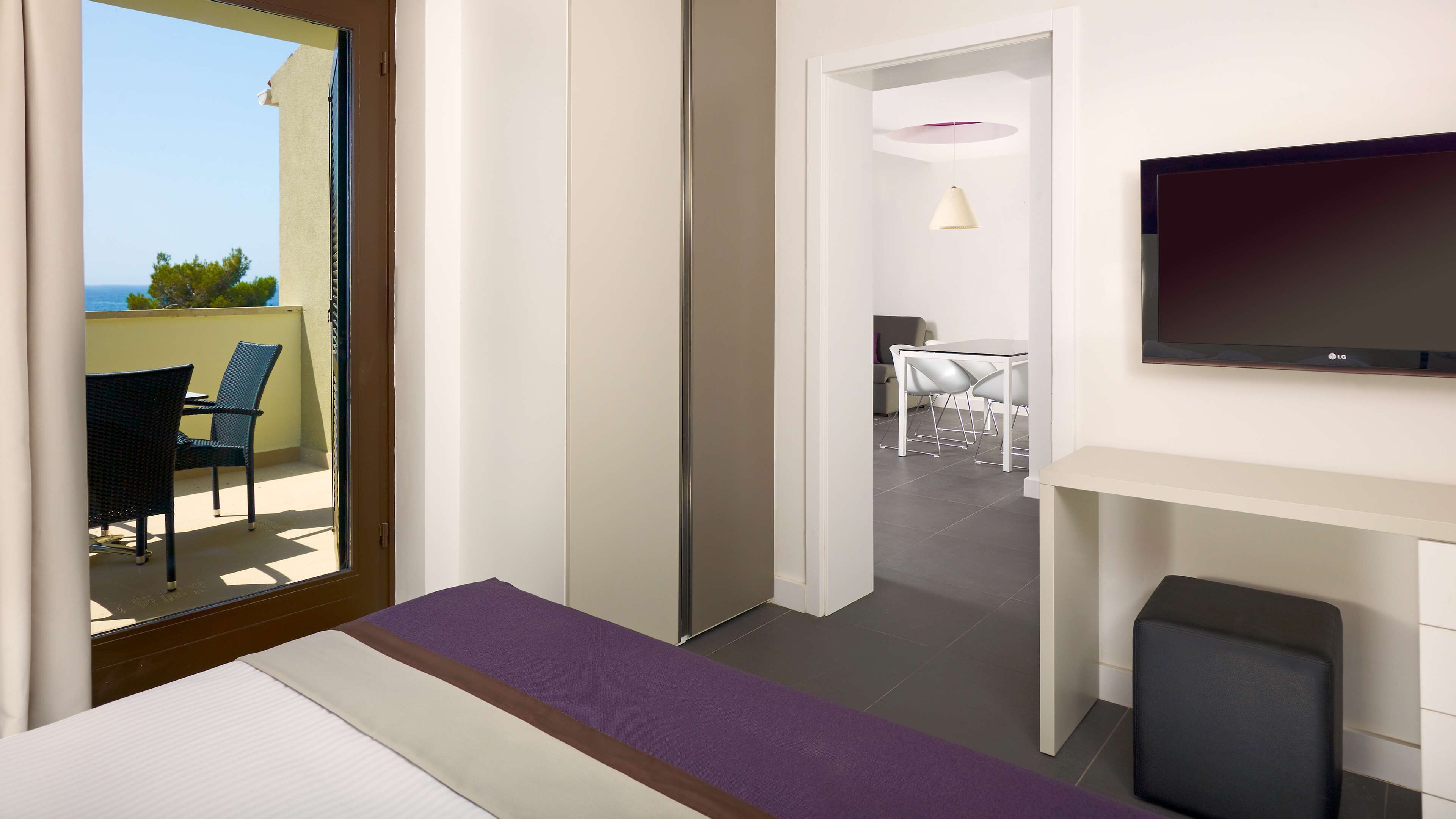 Ein Hotelzimmer mit Bett, Fernseher, Balkon mit Stühlen und einem Essbereich mit Tisch und Stühlen.