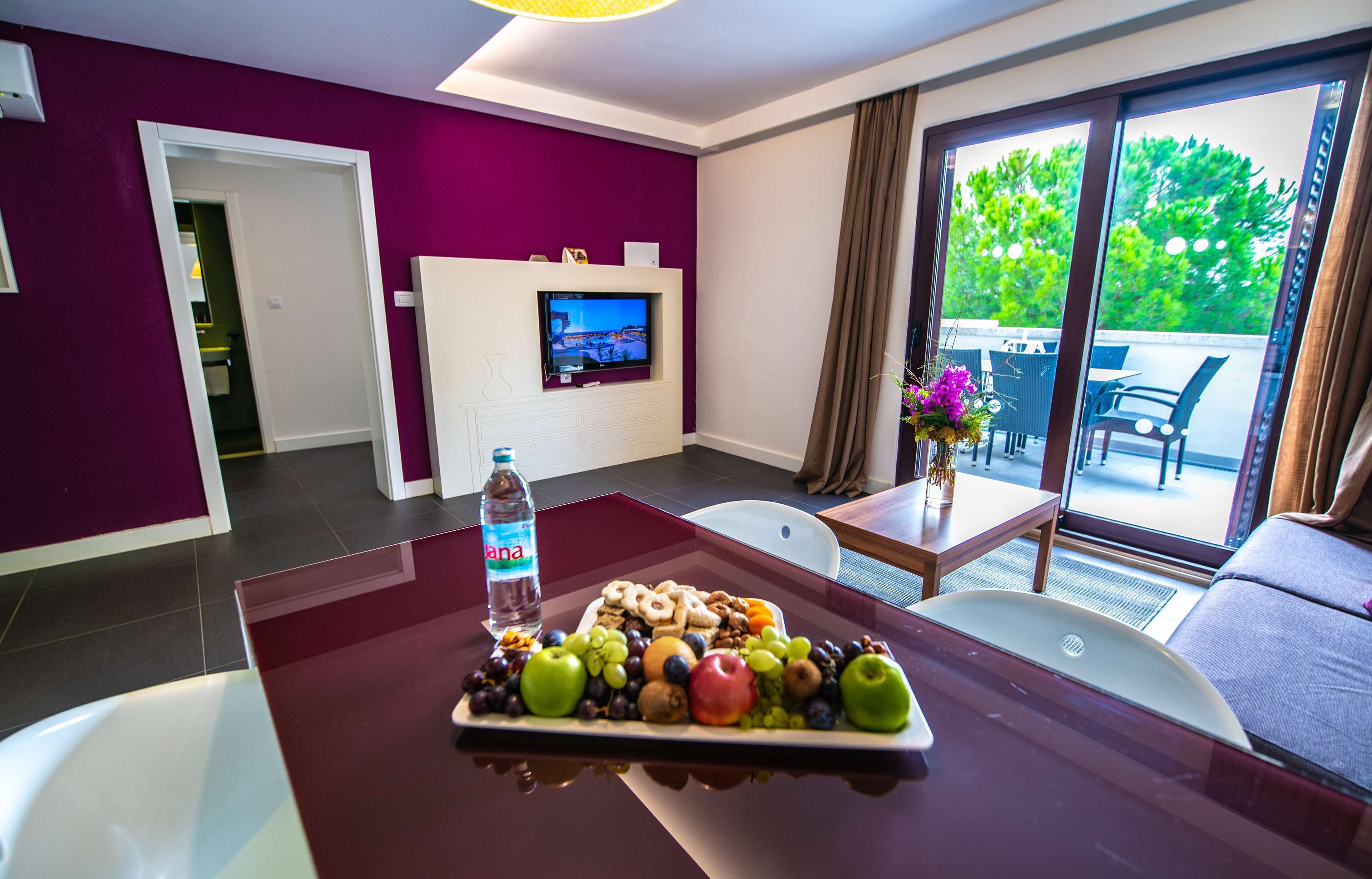 A modern hotel room with purple walls, a flat-screen TV, a dining table with fruit and nuts, and a balcony with outdoor furniture.