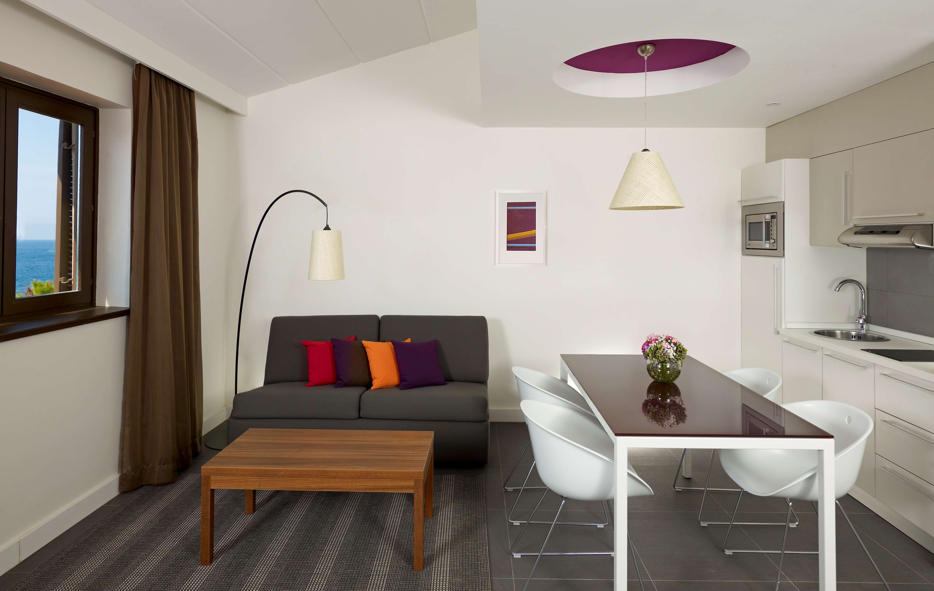 A modern living room with a sofa, coffee table, and dining table. There is a kitchen area with a microwave and sink.
