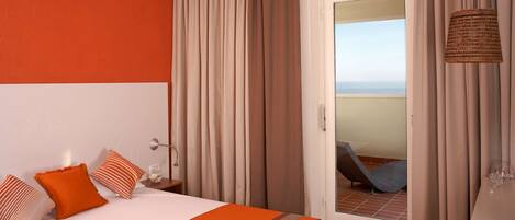 A hotel room with a bed, a nightstand, a lamp, and a view of the sea through a door.