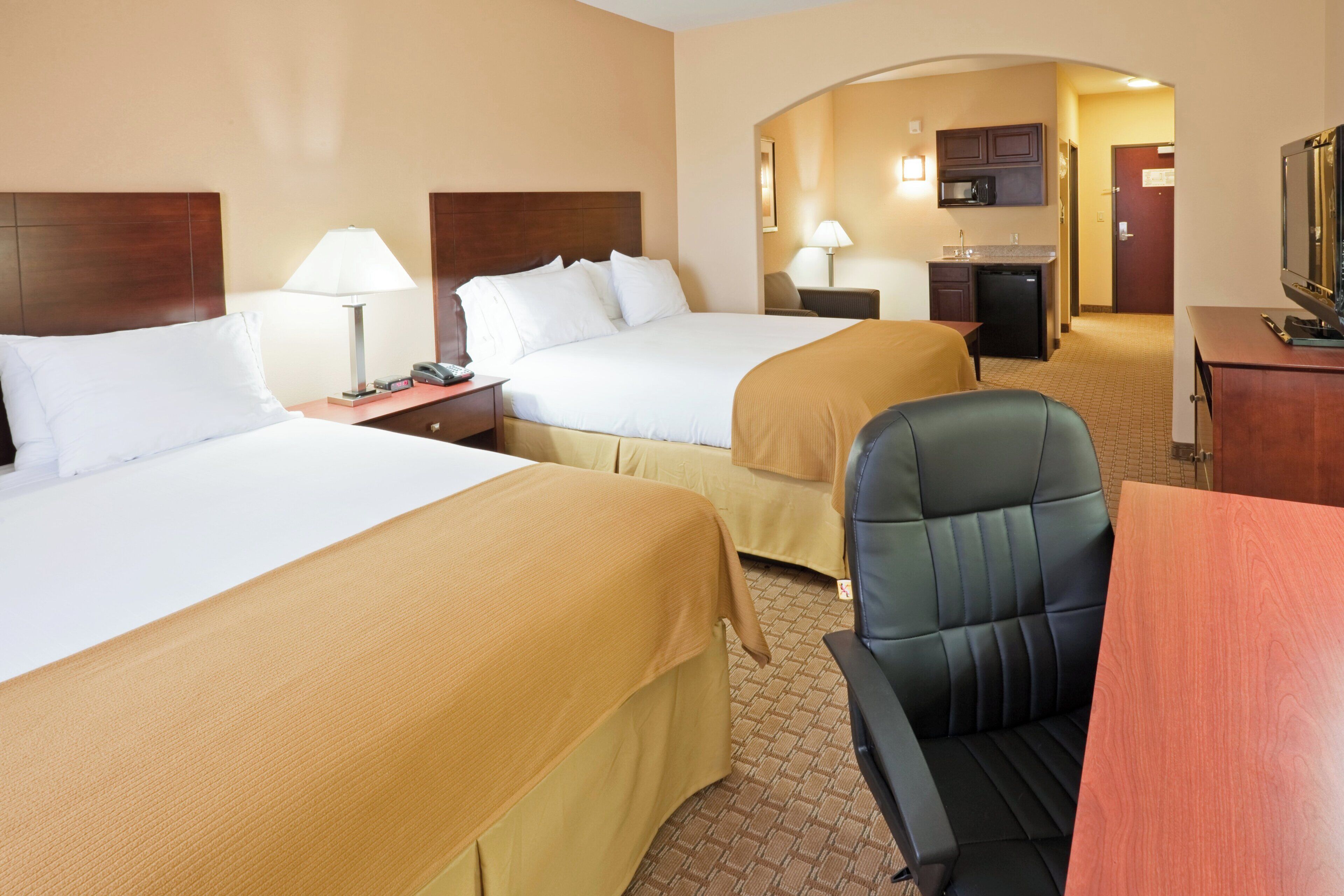 A hotel room with two beds, a desk, a chair, a small kitchenette, and a television.