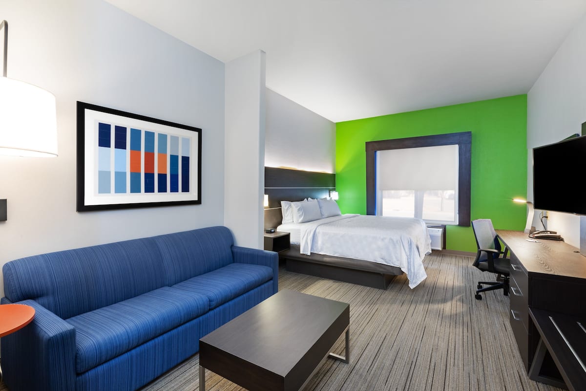 A hotel room with a blue sofa, a bed with white bedding, a desk with a chair, and a large window.