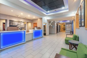 Un vestíbulo de hotel con mostrador de recepción, un área de descanso con sillas verdes y un pasillo que conduce a otras habitaciones.