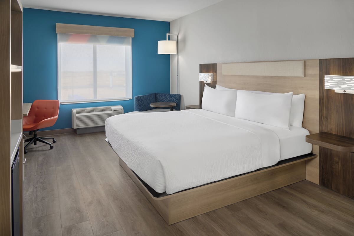 A modern hotel room with a large bed, a desk, a chair, and a window with blinds.