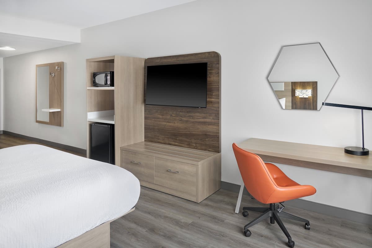A modern hotel room with a bed, a wooden dresser, a desk with a chair, a flat-screen TV, a mirror, and a microwave.