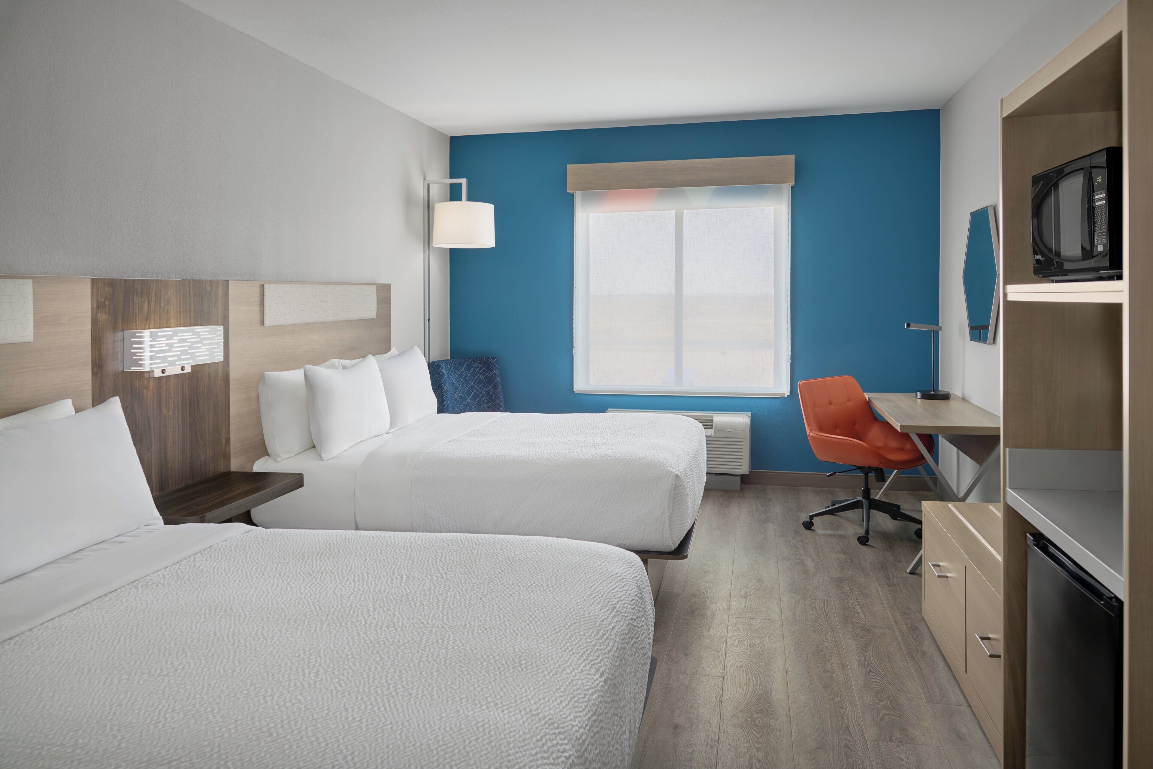 A hotel room with two beds, a desk, an orange chair, a microwave, and a large window.