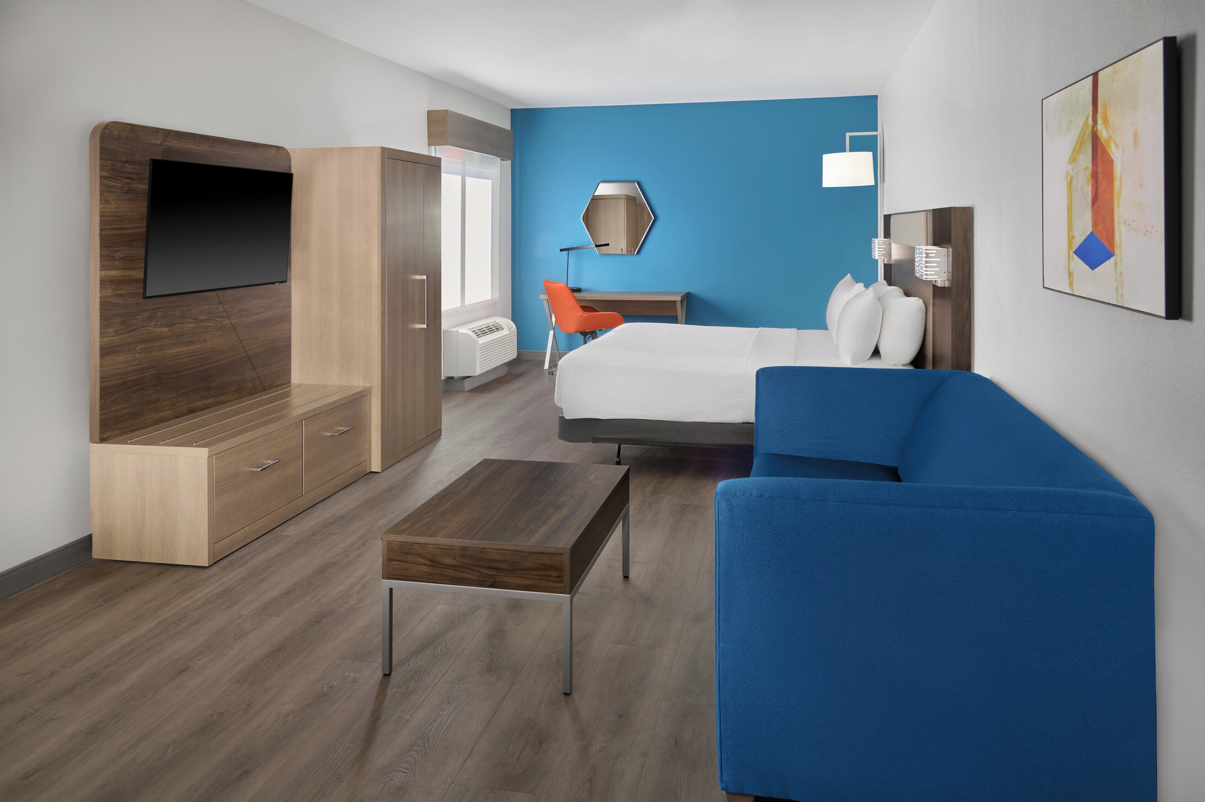 A modern hotel room with a bed, a blue sofa, a wooden TV stand, a desk, and a chair.