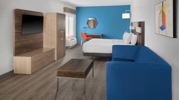 A modern hotel room with a bed, a blue sofa, a wooden TV stand, a desk, and a chair.