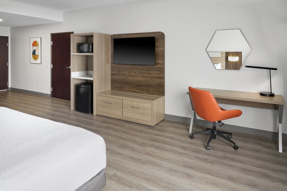 A modern bedroom with a bed, a desk, an orange chair, a wardrobe, a TV, and a mirror.