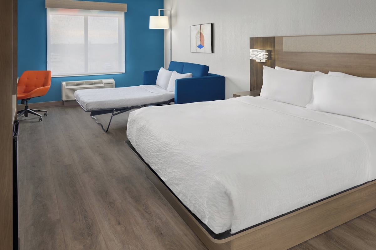 A hotel room with a large bed, a blue armchair, a small desk, and a window with blinds.