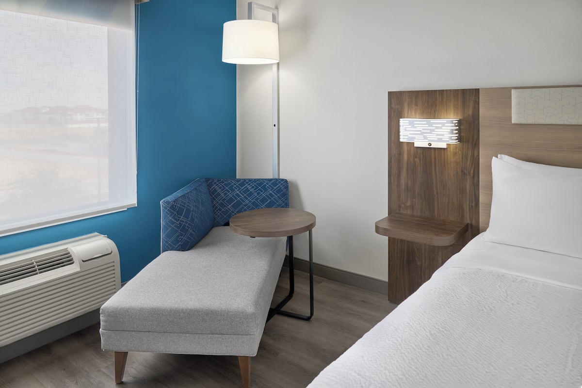 A hotel room with a bed, a bench, a small table, a wall-mounted lamp, and a heater.