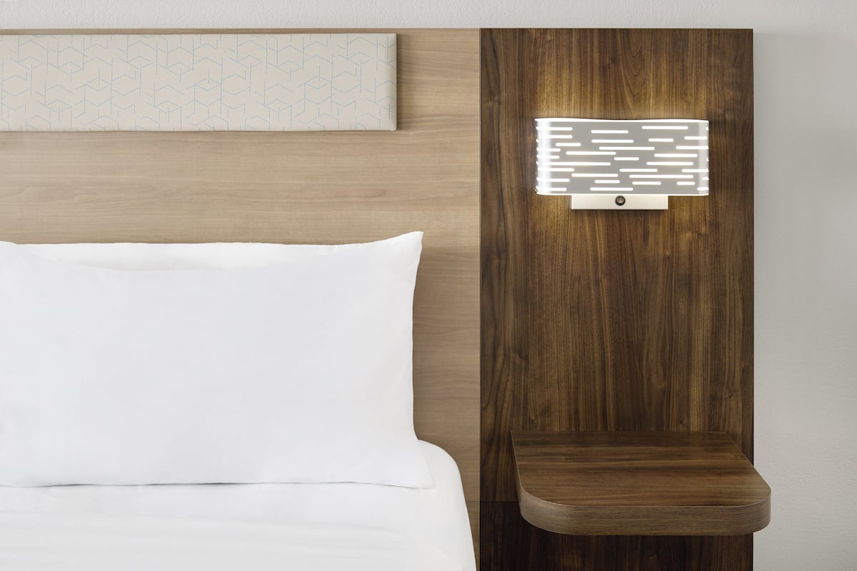 A modern hotel room with a wooden headboard, a bedside table, and a wall-mounted light fixture.