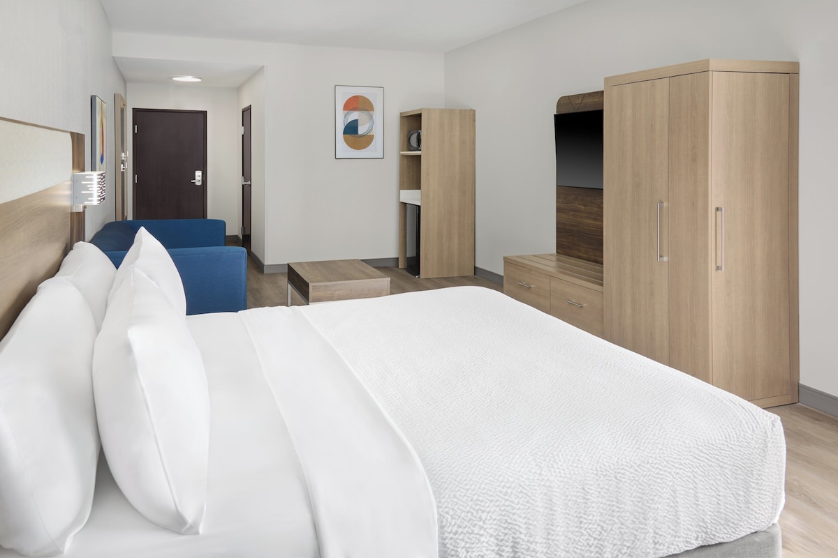 A hotel room with a large bed, a desk, a chair, a wardrobe, and a small table.
