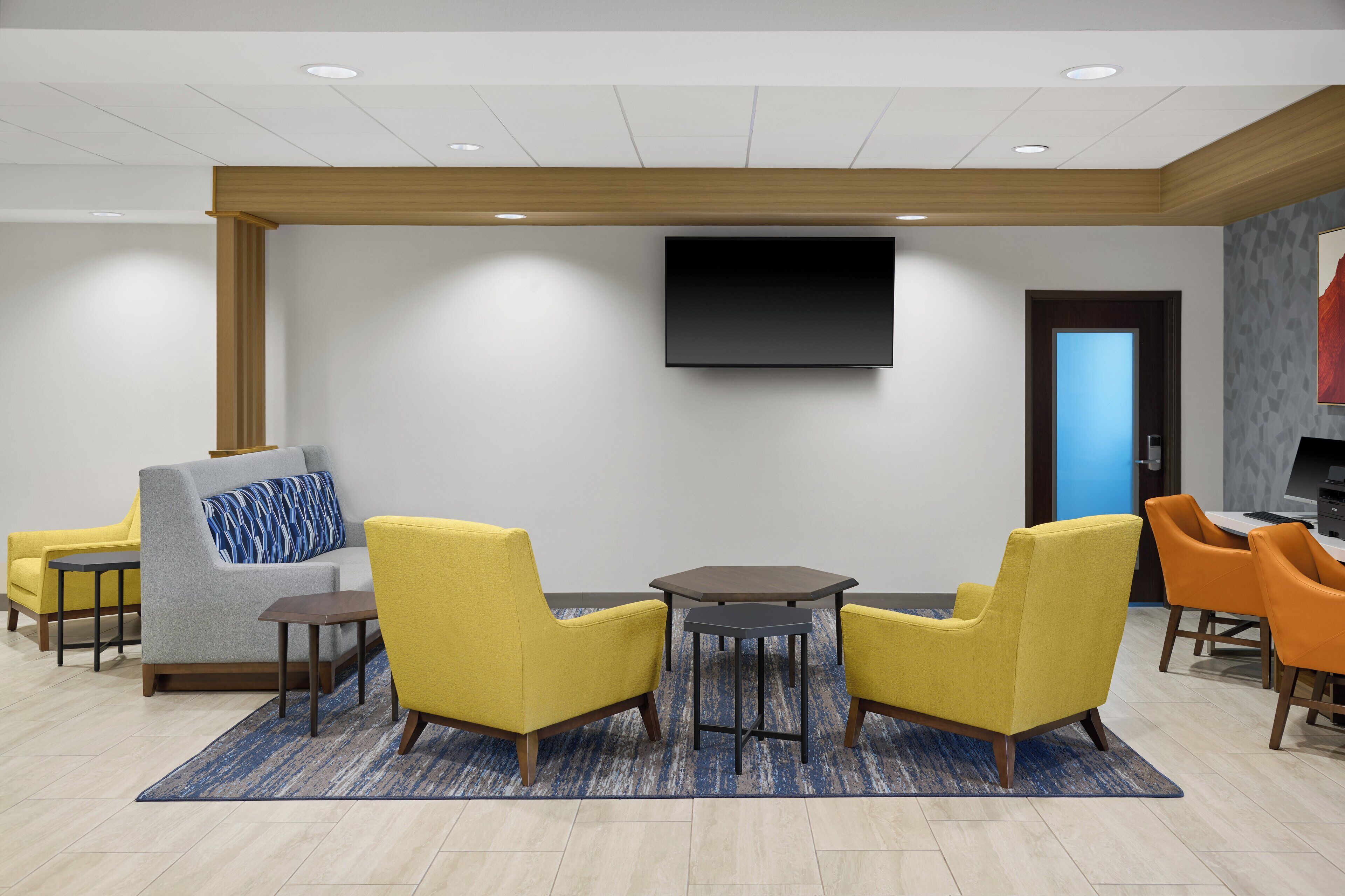 A modern lobby with a flat-screen TV, yellow and grey armchairs, a small round table, and a blue patterned rug.
