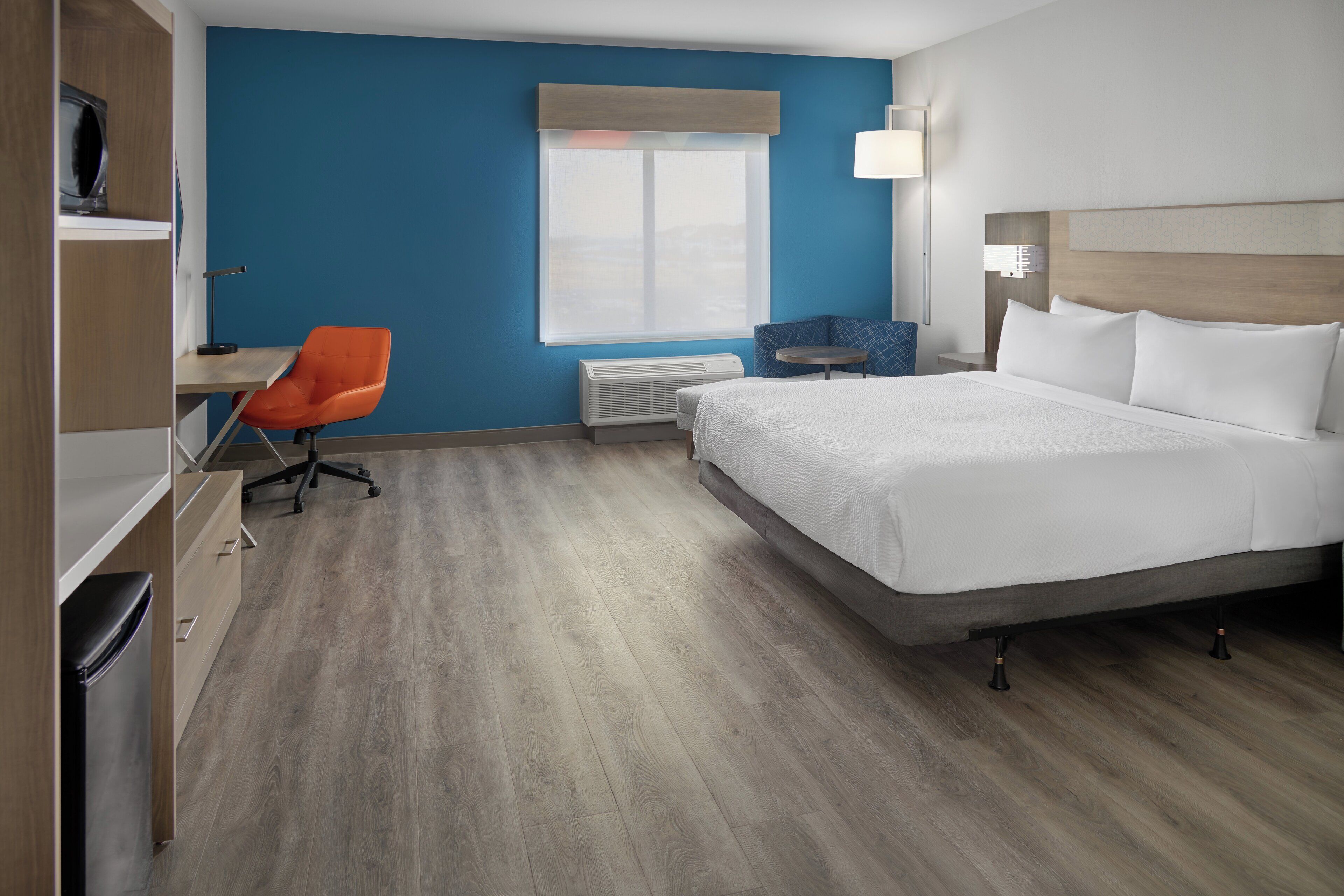 A hotel room with a bed, a desk, an orange chair, a window, and a wall-mounted lamp.