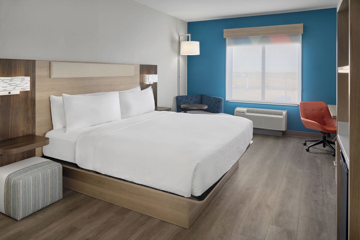 A modern hotel room with a large bed, a desk, a chair, and a window with blinds.