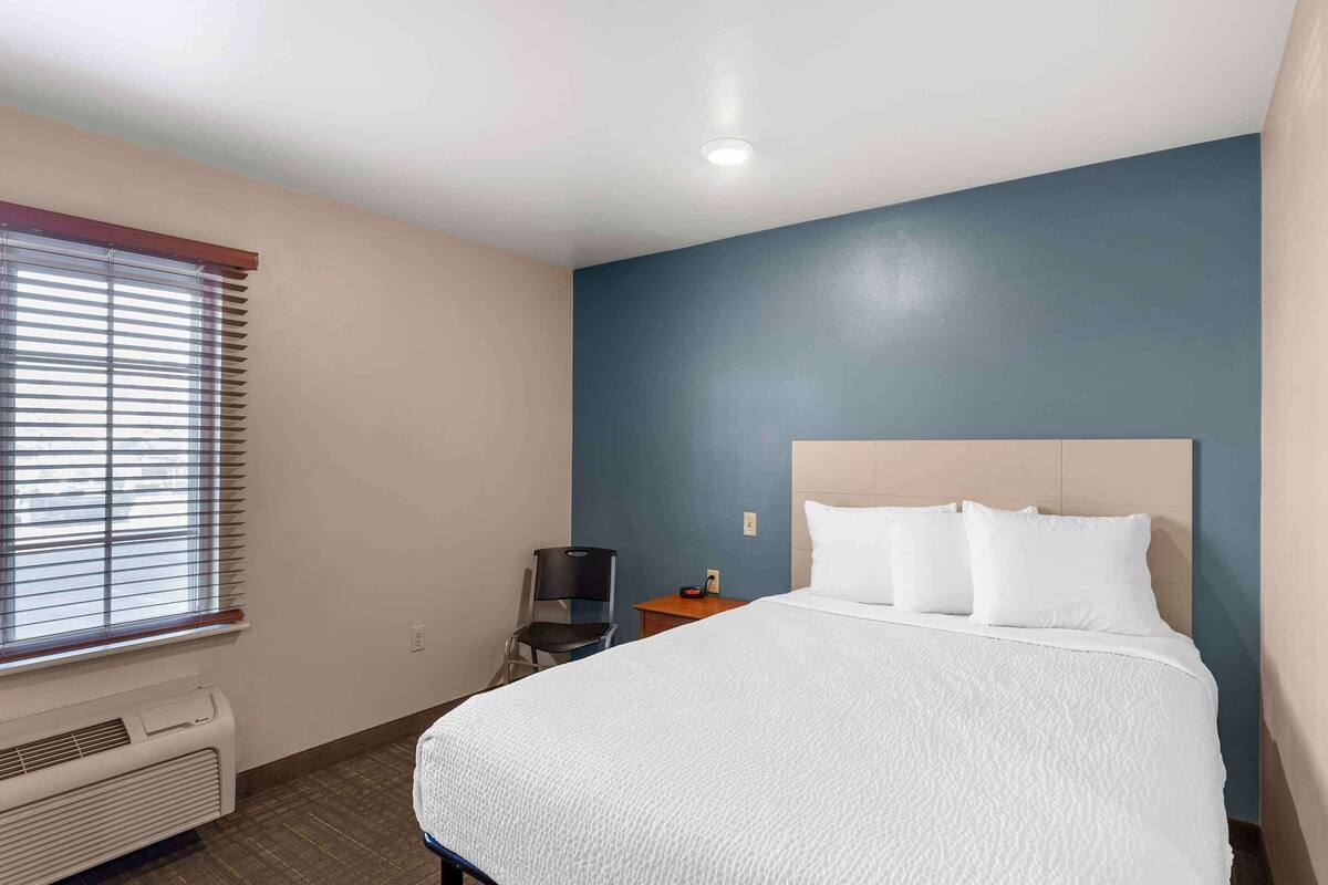 Una habitación de hotel con cama, una silla, una mesita y una ventana con persianas.