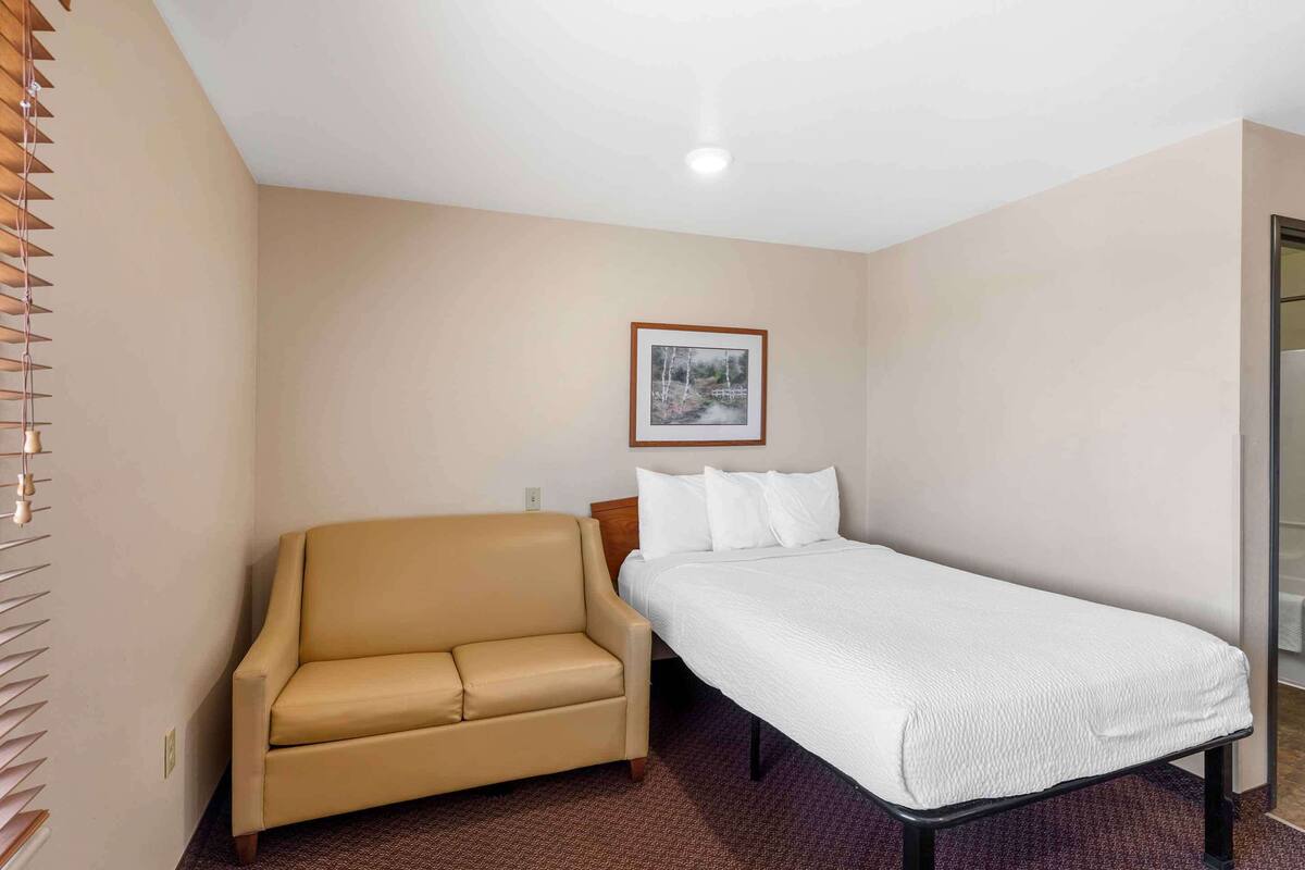 Una cama individual con colchón y almohadas blancos, un sofá color beige, un cuadro en la pared y una puerta que da a un baño.