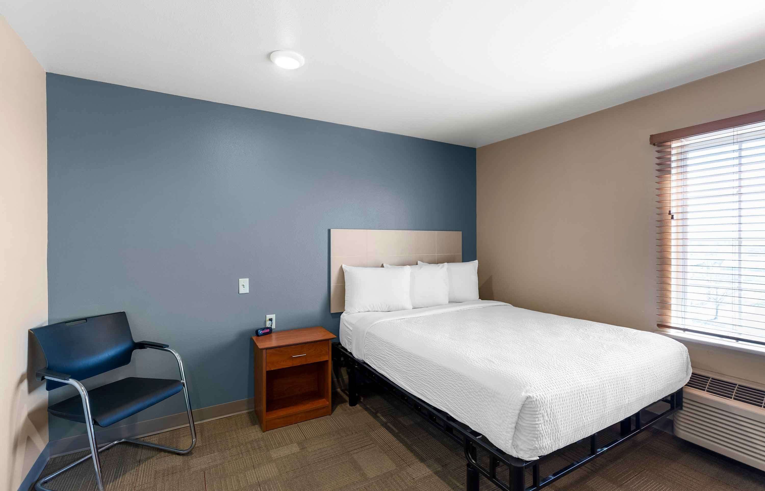 Une chambre d’hôtel avec un lit, une chaise, une table de chevet et une fenêtre avec des stores.