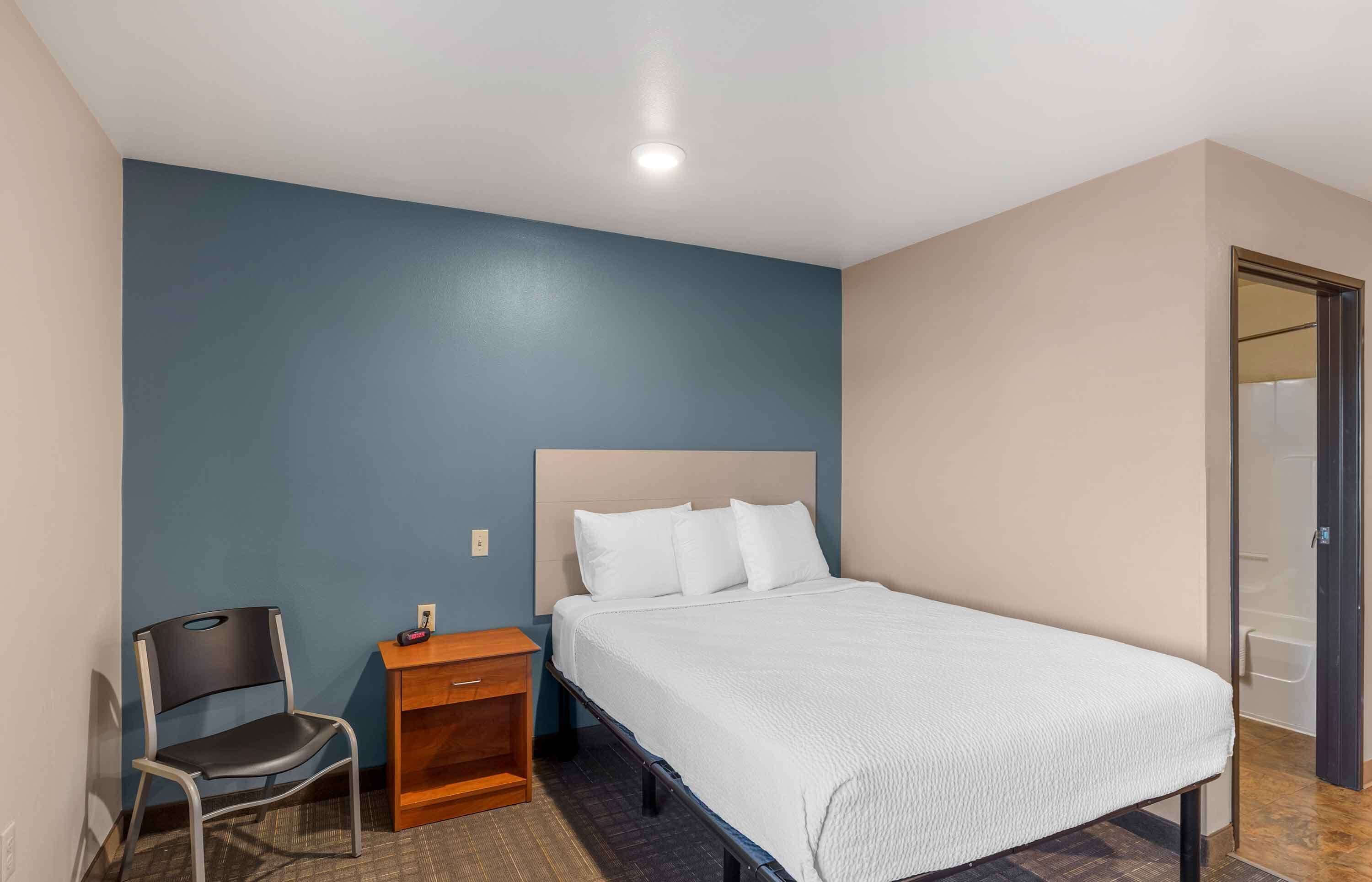A bedroom with a bed, a chair, a nightstand, and a door leading to a bathroom.