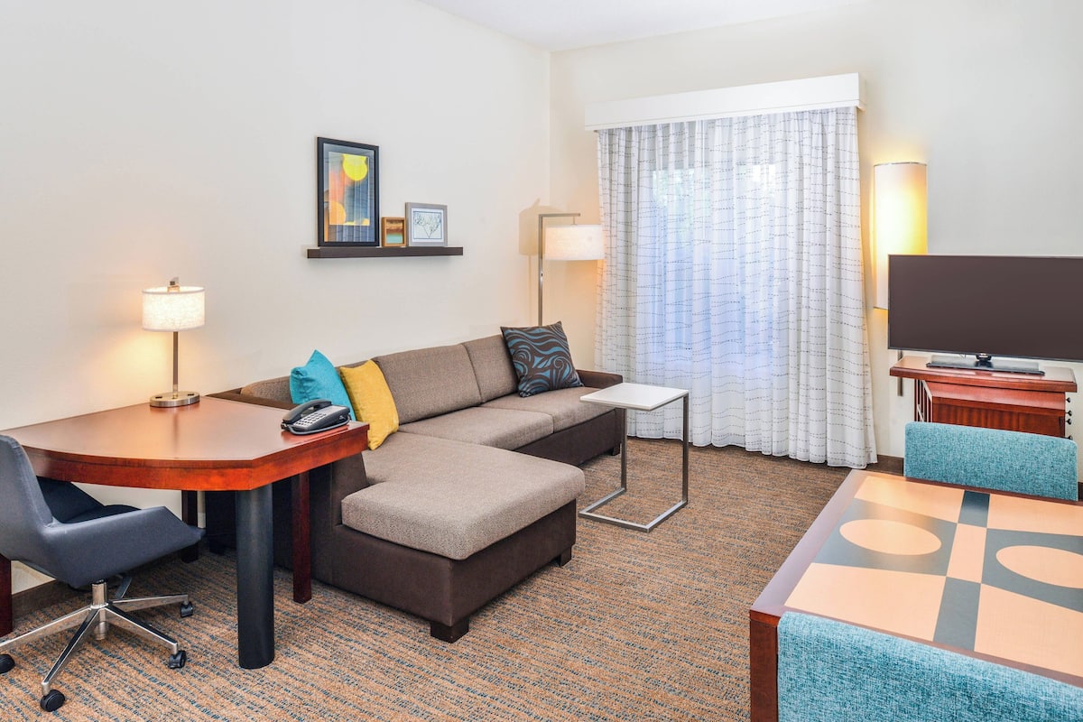 A hotel room with a sofa, a desk, a chair, a TV, and a lamp.