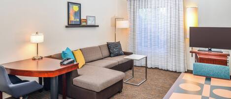 A hotel room with a sofa, a desk, a chair, a TV, and a lamp.