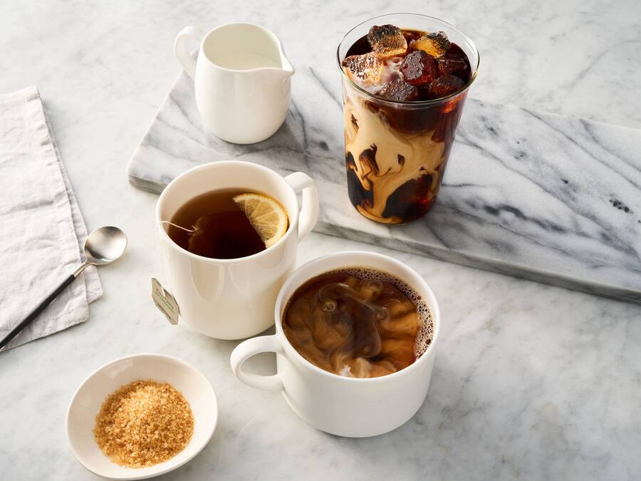 Une table en marbre sur laquelle reposent deux tasses de thé blanches, dont l’une contient une tranche de citron, un verre de thé glacé avec des glaçons, un petit bol de sucre et un pichet blanc.