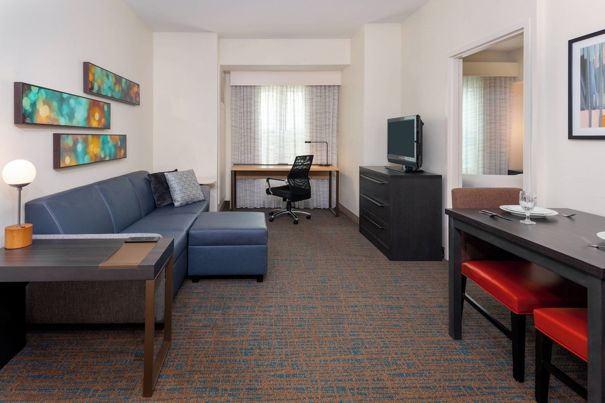 A hotel room with a sofa, a desk with a chair, a television, and artwork on the walls.