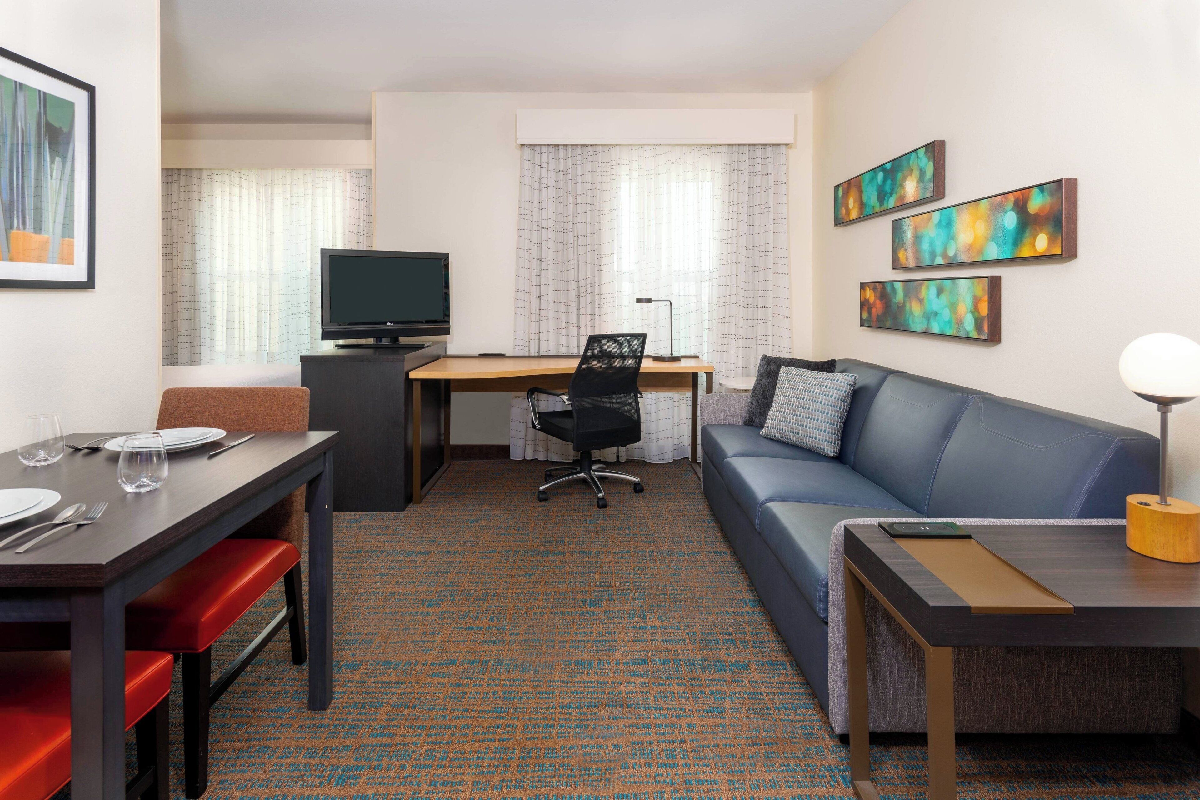 A hotel room with a dining table, a sofa, a TV, and artwork on the walls.