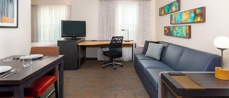 A hotel room with a dining table, a sofa, a TV, and artwork on the walls.
