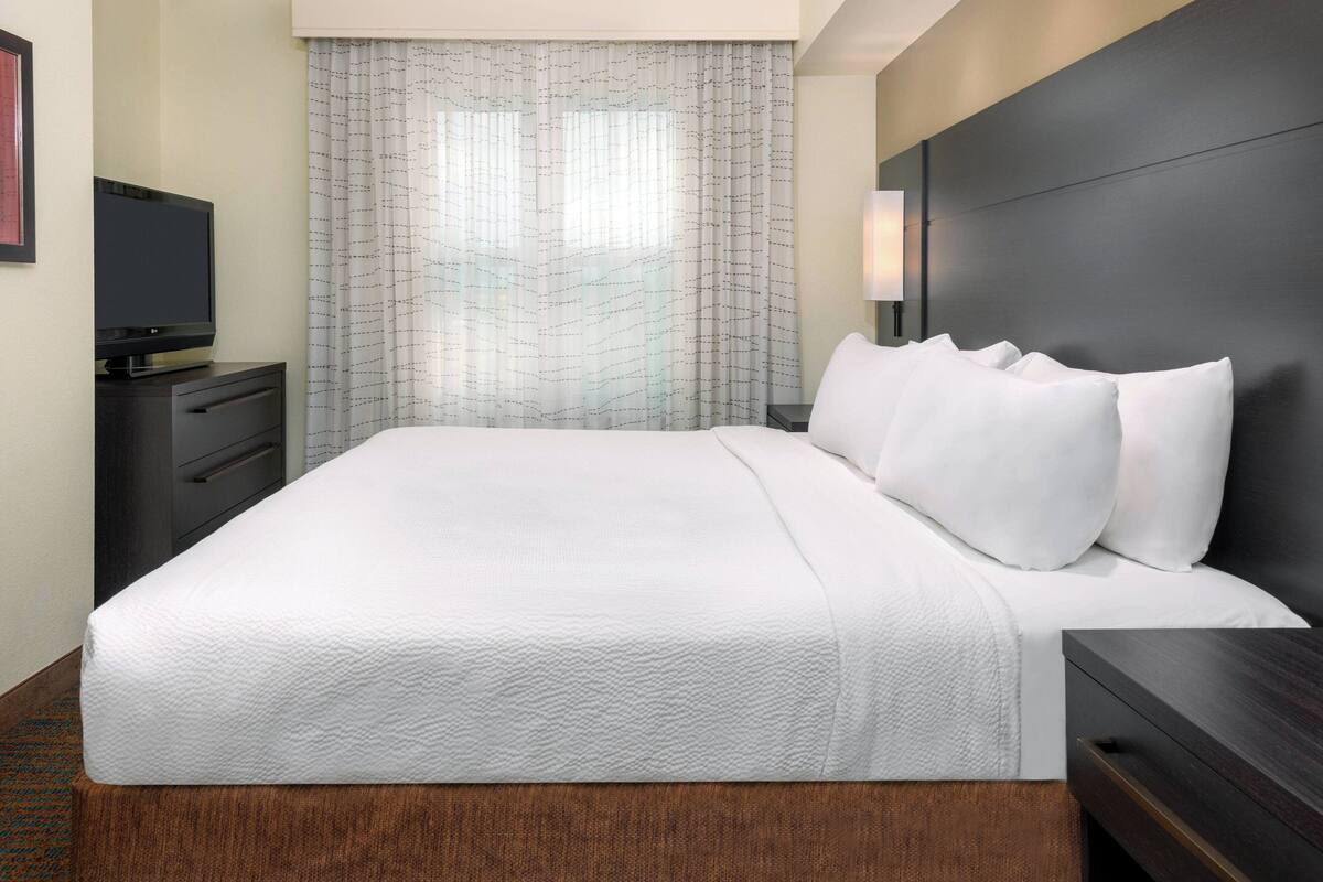 A hotel room with a large bed, a flat-screen TV on a dresser, and a window with sheer curtains.