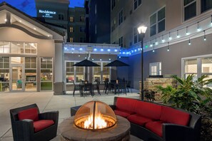 Un área de patio al aire libre con un hogar para fuego, muebles de mimbre rojo y negro, y luces colgantes.