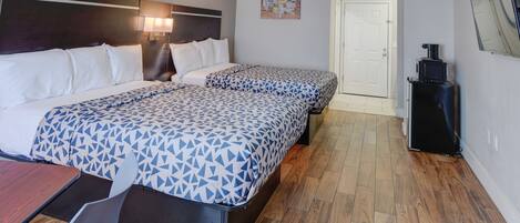 Ein Hotelzimmer mit zwei Betten, einem Schreibtisch und einem Sessel. Eine Tür führt in einen weiteren Raum, und ein kleiner Tisch mit einer Kaffeemaschine steht bereit.