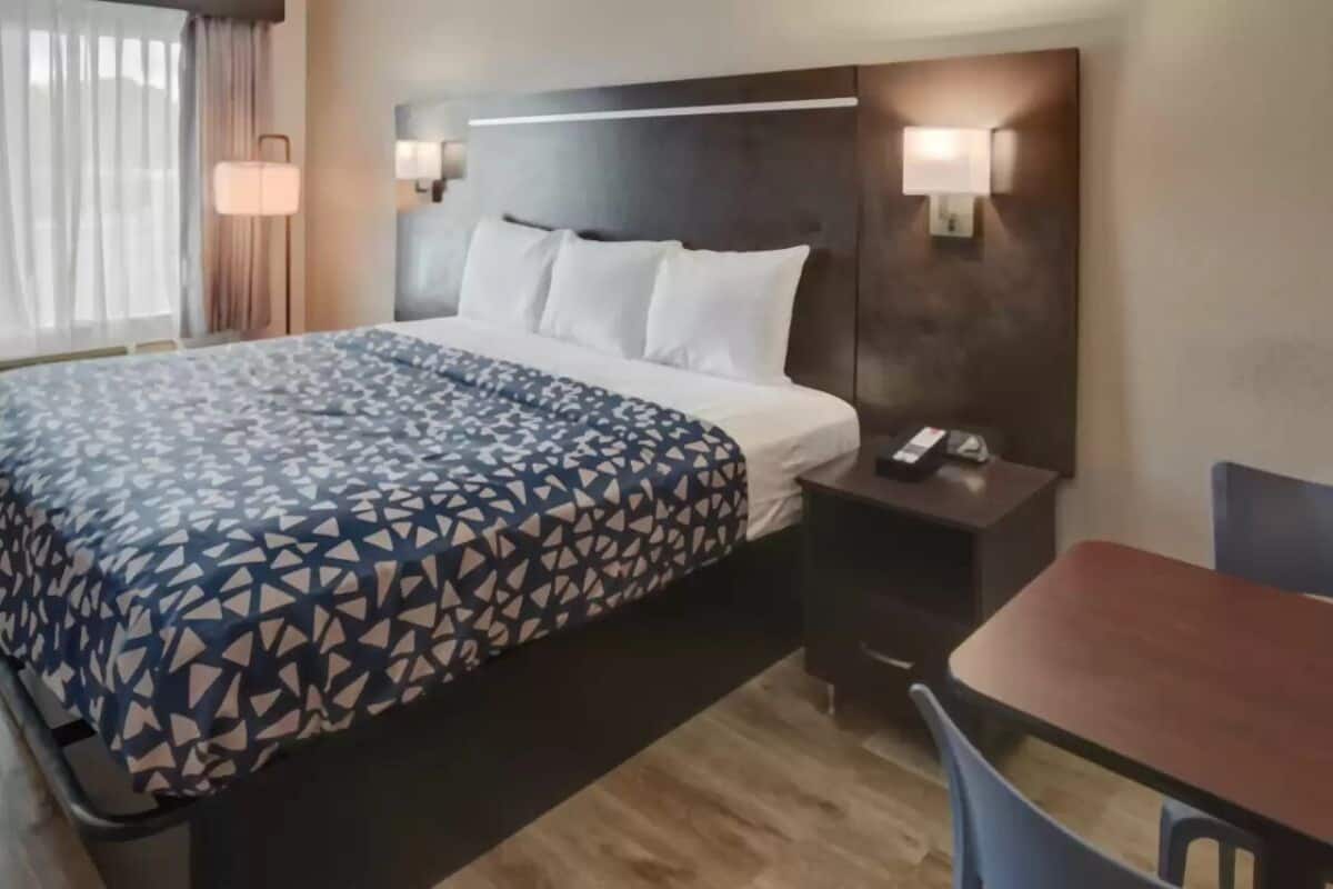 Habitación de hotel con cama, mesita de noche, lámpara y una pequeña mesa con sillas.