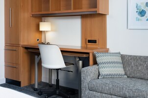 Un espacio de oficina moderno con un escritorio de madera, una silla de oficina blanca, un sofá gris con un cojín estampado y una lámpara fijada a la pared.