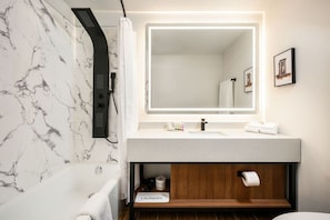 A bathroom with a marble wall, a shower, a sink with a mirror, and a wooden vanity. - Acclaim Hotel By CLIQUE (Calgary)