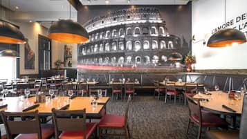 A restaurant interior with multiple dining tables and booths, a large colosseum mural on the wall, and pendant lighting.