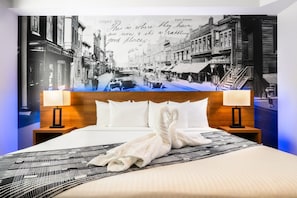 A hotel room with a bed, two bedside tables, a headboard, and a large wall mural of a street scene. - Acclaim Hotel By CLIQUE (Calgary)