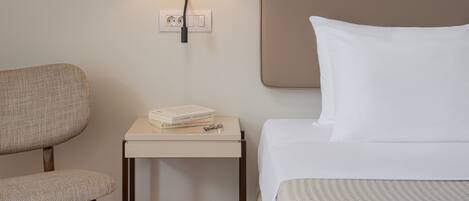 A hotel room with a bed, a chair, a small table with a book, a wall-mounted lamp, and a power outlet.